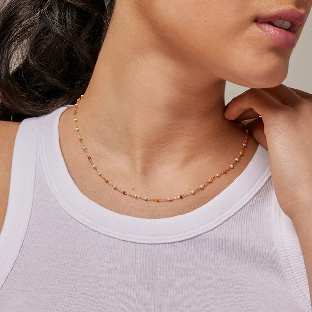 A close-up of a model wearing a delicate beaded necklace with small gold and orange-red beads, paired with a white ribbed tank top.
