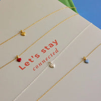 Four delicate chain necklaces with small heart charms in gold, red, silver, and light blue, arranged diagonally on a beige card with the text 'Let's stay connected'.