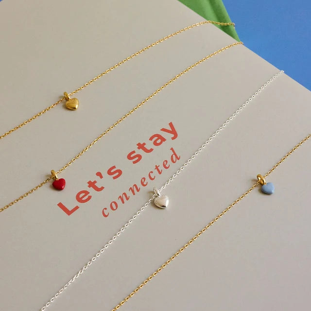 Four delicate chain necklaces with small heart charms in gold, red, silver, and light blue, arranged diagonally on a beige card with the text 'Let's stay connected'.