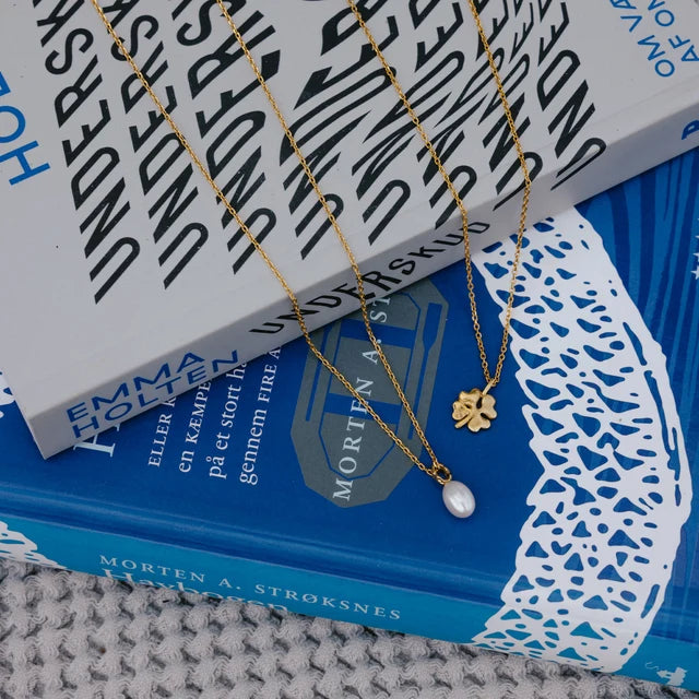 Two delicate gold necklaces displayed on top of books. One necklace has a small gold clover charm, and the other has a teardrop-shaped white pearl pendant.
