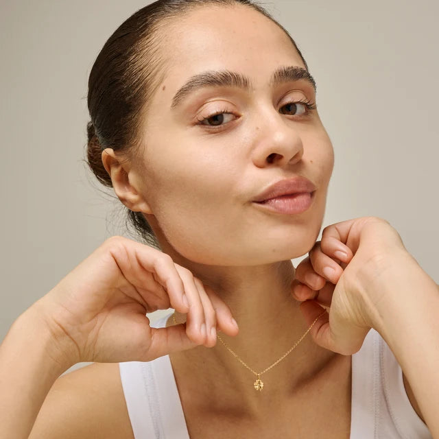 A close-up lifestyle shot of a model wearing a delicate gold necklace with a small clover pendant against a neutral beige background.