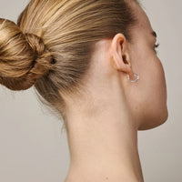 A close-up side profile of a woman with blonde hair in a bun, wearing a small silver heart-shaped hoop earring against a plain light gray background.
