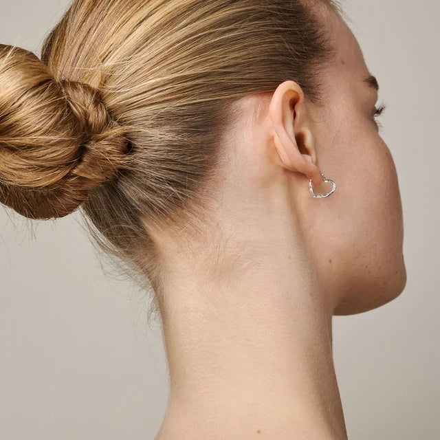 A close-up side profile of a woman with blonde hair in a bun, wearing a small silver heart-shaped hoop earring against a plain light gray background.
