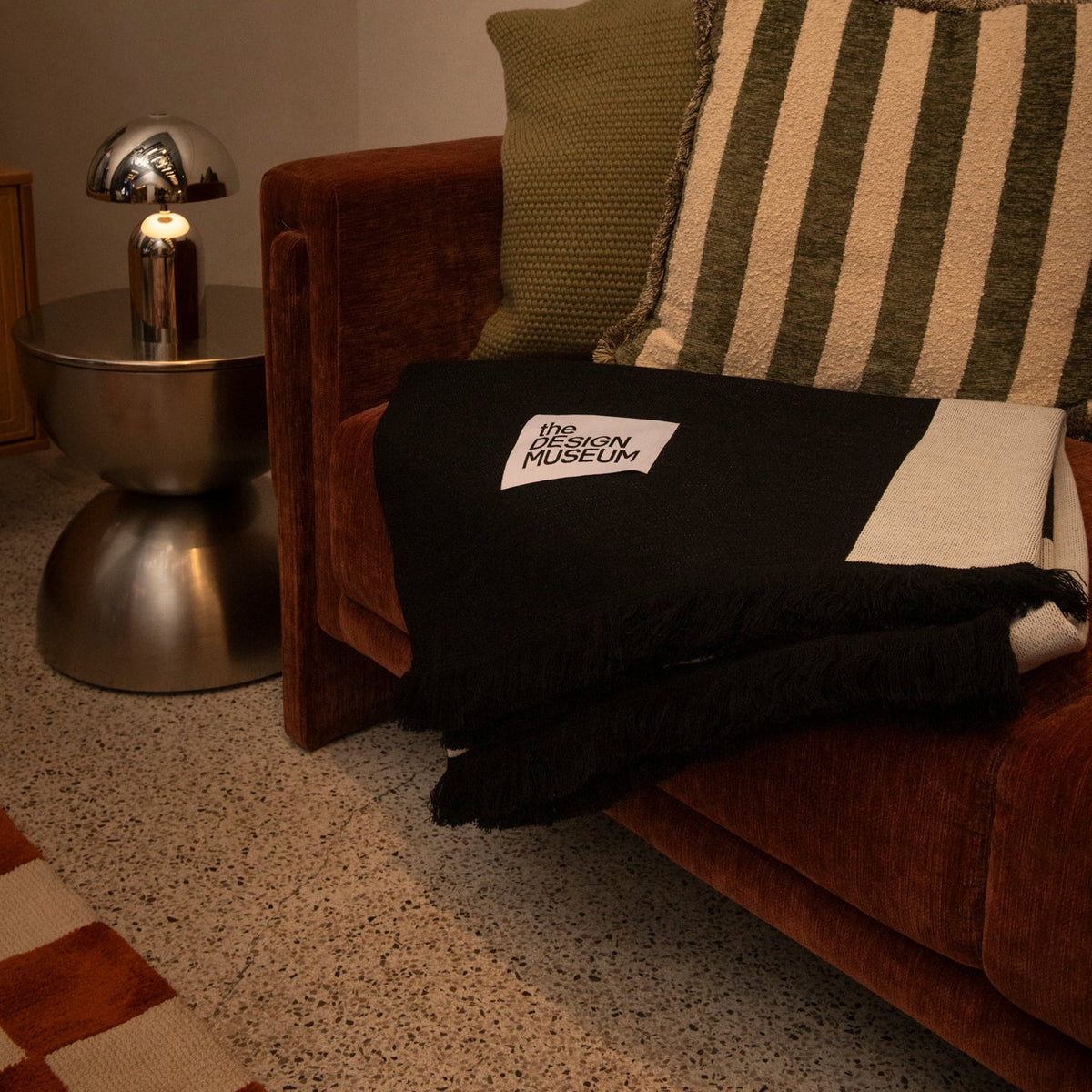 A black throw blanket with a white 'THE NORTH FACE' logo patch is draped over a brown velvet sofa next to a striped green and cream pillow, in a warmly lit room.