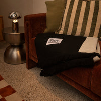 A black throw blanket with a white 'THE NORTH FACE' logo patch is draped over a brown velvet sofa next to a striped green and cream pillow, in a warmly lit room.