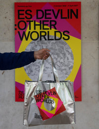 A person holds a metallic silver tote bag in front of a colorful poster. The tote bag has a geometric design in pink, yellow, and red with black text reading 'ES DEVLIN : OTHER WORLDS' and 'The Design Museum.