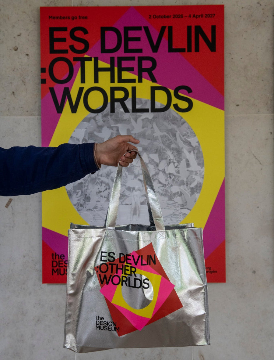 A person holds a metallic silver tote bag in front of a colorful poster. The tote bag has a geometric design in pink, yellow, and red with black text reading 'ES DEVLIN : OTHER WORLDS' and 'The Design Museum.