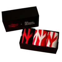 Three folded knit socks in a black presentation box, with a matching black box behind them on a red background. The socks have large abstract red and white Y-shaped knit patterns, and the front box is labeled 'the DESIGN MUSEUM x' with a red logo.