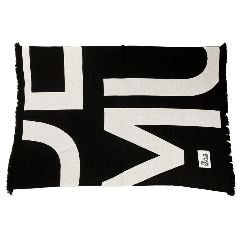 A black and off-white woven throw blanket with fringe on the edges, featuring a large abstract geometric pattern. A small white brand tag is visible in the bottom right corner.