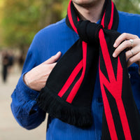 A close-up lifestyle shot of a person wearing a black knit scarf with bold red stripes and fringe, styled over a blue shirt in an outdoor park setting.