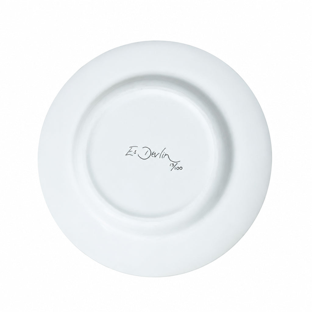 A white ceramic plate photographed from above on a light wood-grain surface, with the printed text 'Es Devlin 700' in the center.