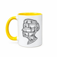 A white ceramic mug with a bright yellow handle and interior, featuring a black line art illustration of a face made of bookshelves on the side, set on a light wood tabletop against a gray concrete wall.