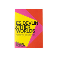 A postcard collection book titled 'ES DEVLIN : OTHER WORLDS' by 'the DESIGN MUSEUM', featuring a bold geometric cover with yellow, pink, and red sections, displayed on a pale yellow background.