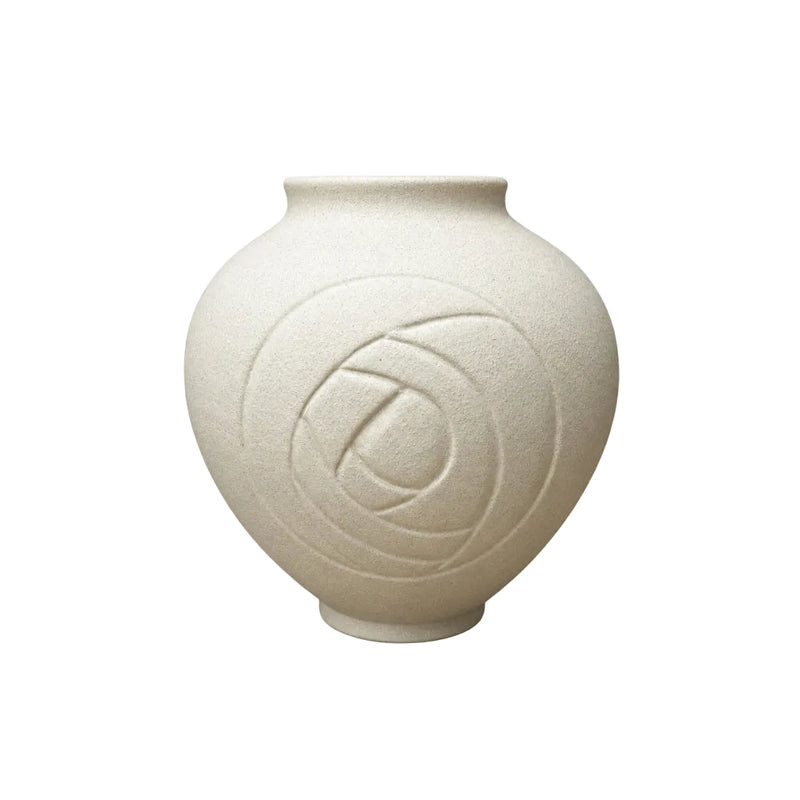 A small matte ceramic vase with a rounded bulbous body, short flared rim, and an embossed circular floral motif on the front, shown on a light wood surface against a gray textured wall.