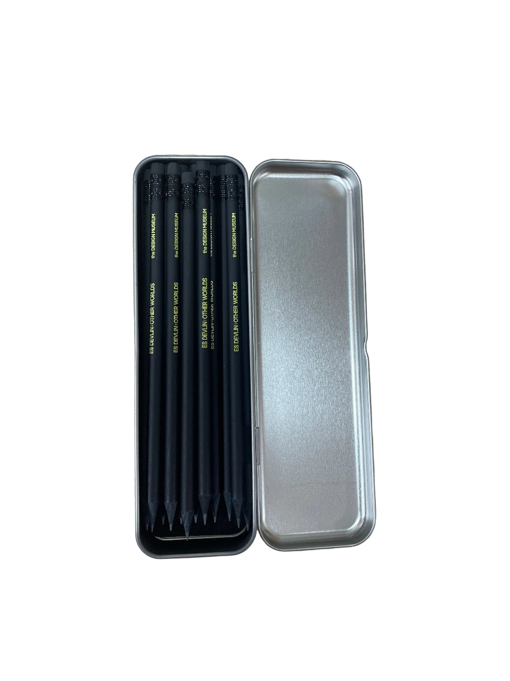 A set of four black pencils with gold lettering inside an open silver metal tin, placed on a light wood grain surface.