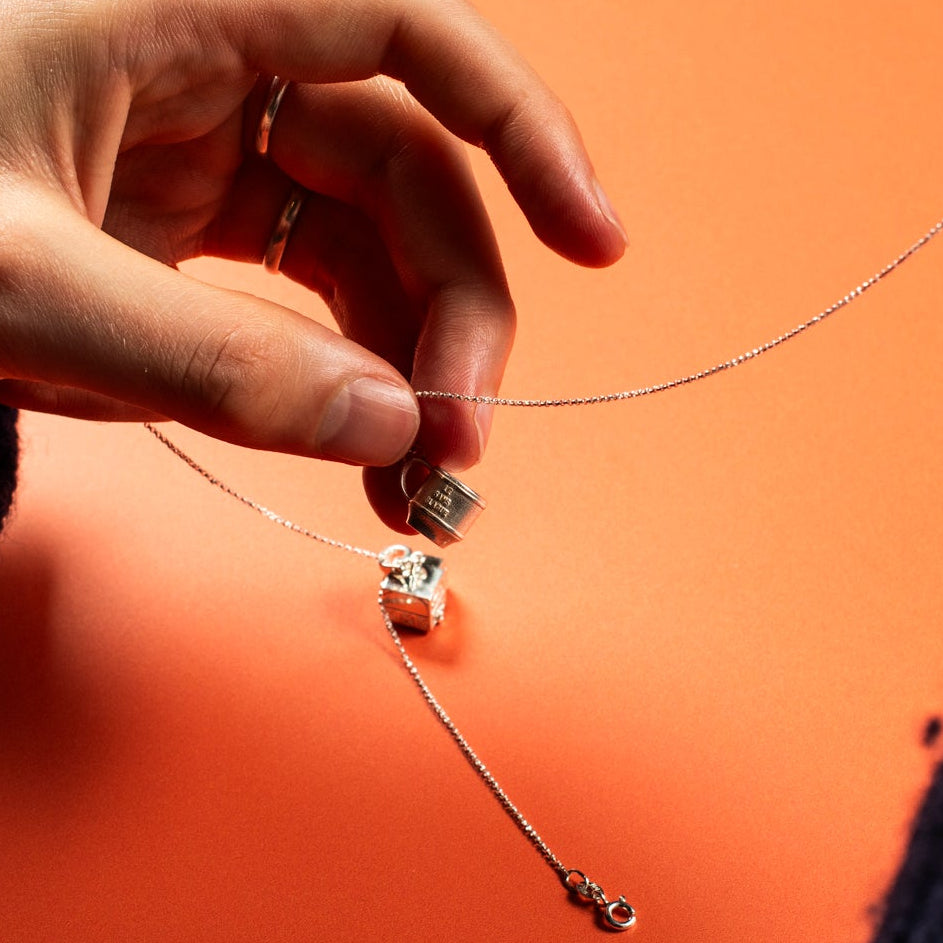 Hands holding a silver necklace with a teacup charm against an orange background