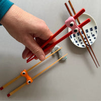 Hand holding red chopsticks with googly eyes on a light gray surface.