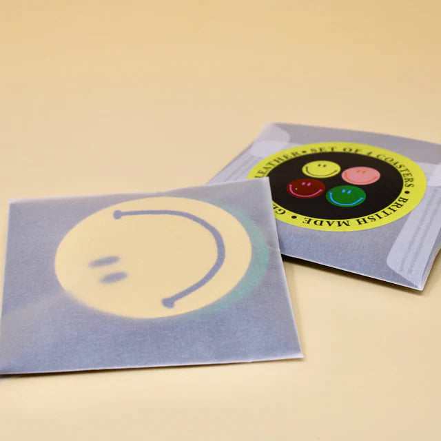 A set of four colourful round coasters with a smiley face design inside semi-translucent paper packaging with a label on the front.