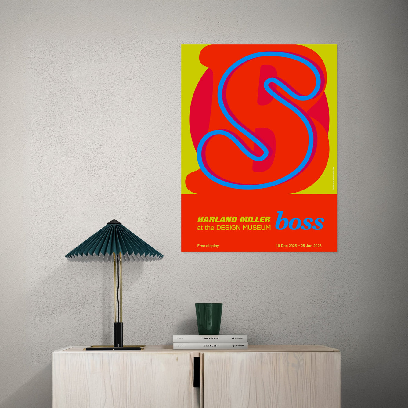 Colorful graphic typography poster with 'boss' design on a gray wall above a wooden cabinet with a lamp and books.