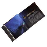 An open printed booklet or magazine spread lying on a tan textured surface, showing a dark fantasy-style illustration of a snowy path through a forest on one page and the title 'THE LONG ROAD' with text columns on the other.