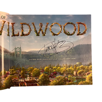 A printed book or magazine page featuring a scenic landscape with large decorative 'Wildwood' lettering across the top, a suspension bridge on the left, and a stylized town scene below. A black handwritten signature is visible near the center-right.