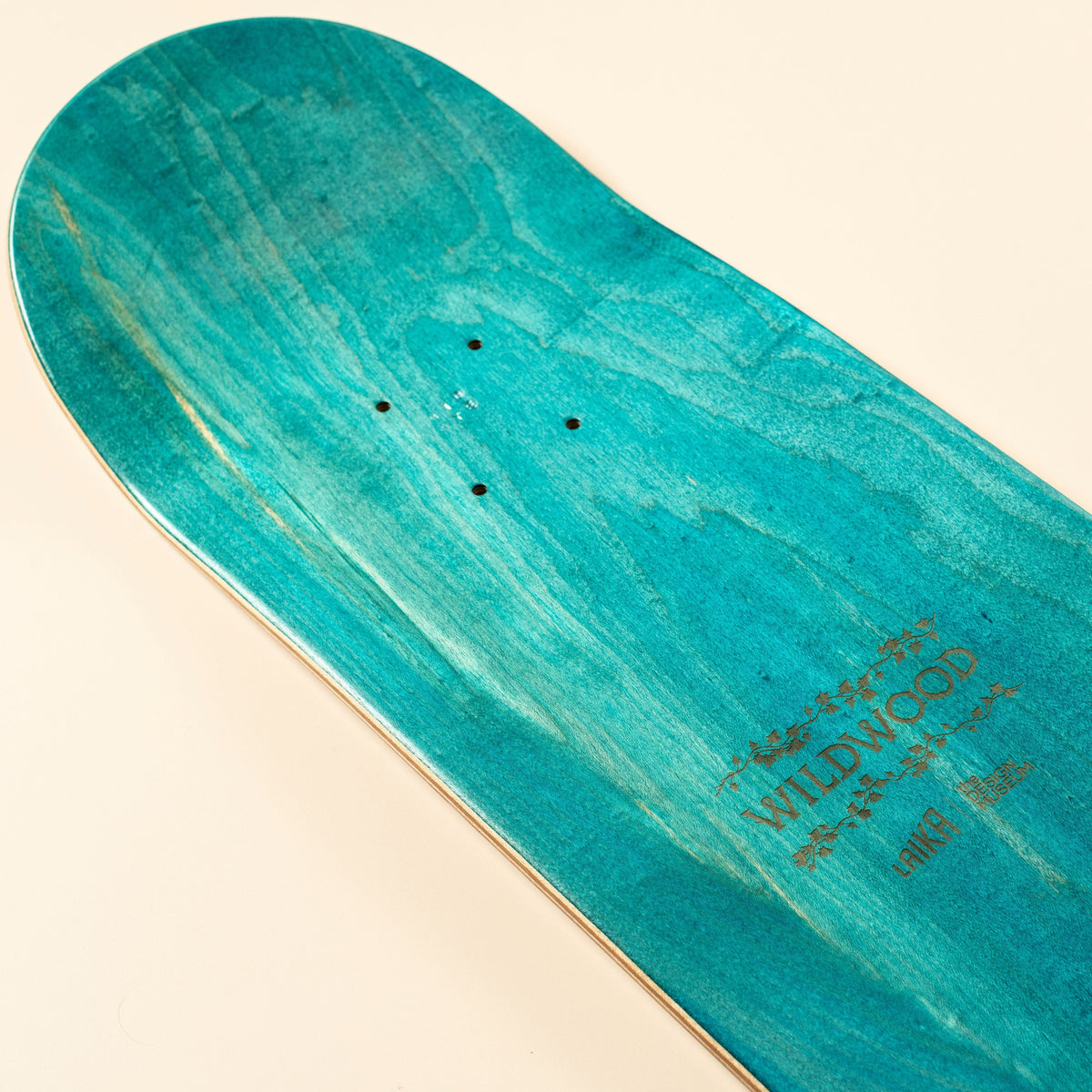 A close-up of a skateboard deck showing the underside with a turquoise and teal wood-grain finish, featuring the brand name 'WILDWOOD' printed in gold near the tail, set against a solid cream background.