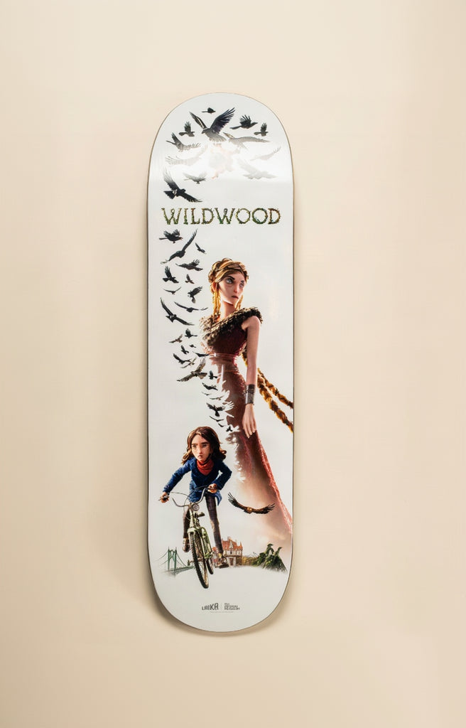 A long skateboard deck with a white base and a detailed graphic print, displayed horizontally against a solid beige background. The graphic features a woman, a child on a bicycle, birds, and the text 'WILDWOOD' and 'windsor rides'.