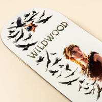 A close-up of a white skateboard deck featuring a printed illustration of a woman with blonde hair, black crows, and the word 'WILDWOOD' in textured green letters, photographed on a warm beige background.