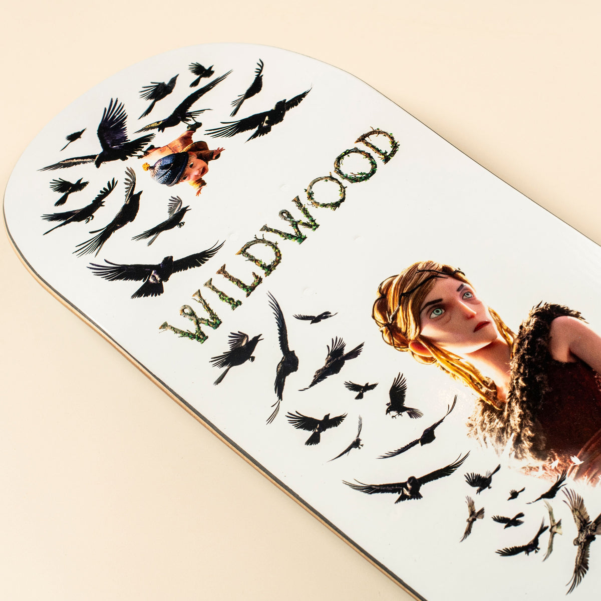 A close-up of a white skateboard deck featuring a printed illustration of a woman with blonde hair, black crows, and the word 'WILDWOOD' in textured green letters, photographed on a warm beige background.