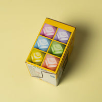 A yellow cardboard box containing six small pastel-colored cube-shaped items arranged in a 2x3 grid, photographed on a plain warm yellow background.