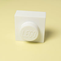 A white LEGO 1x1 brick standing upright on a pale yellow background, with the embossed LEGO logo visible on the top circular face.