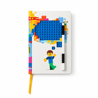 A notebook with a LEGO-inspired design, featuring a blue brick-pattern shape, a small LEGO minifigure, and a yellow ribbon bookmark, photographed on a pale yellow background.