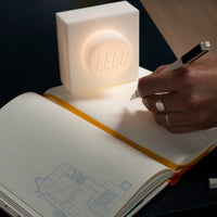 A hand holding a pen writes in an open notebook next to a glowing white LEGO brick-shaped lamp on a dark table.
