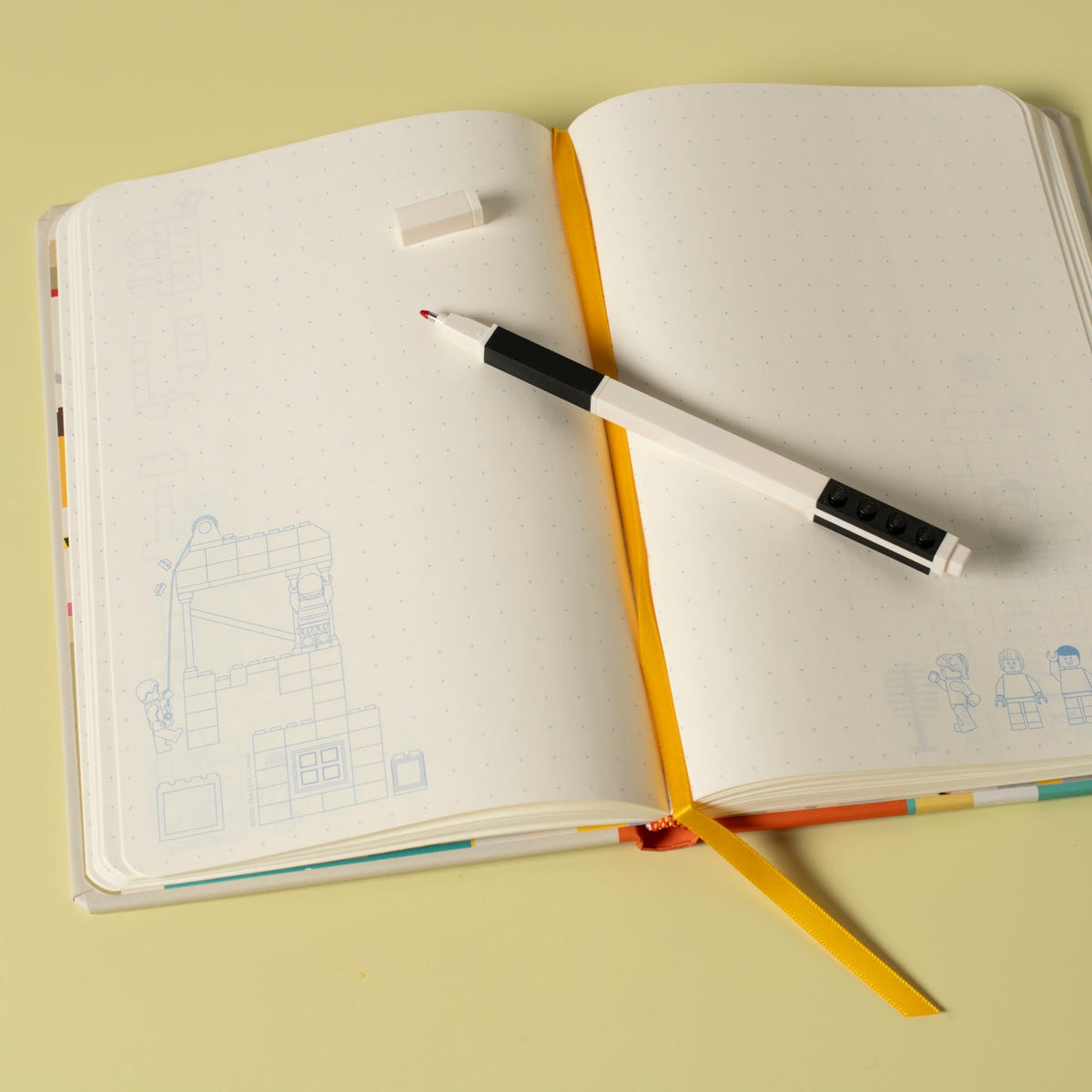 An open notebook with blank white pages, a yellow ribbon bookmark, and a black-and-white pen resting across the spread, photographed on a pale yellow background.