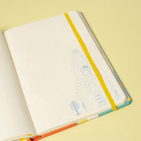 An open notebook with blank white pages, a yellow elastic closure, and a colorful striped cover, photographed on a pale yellow background.
