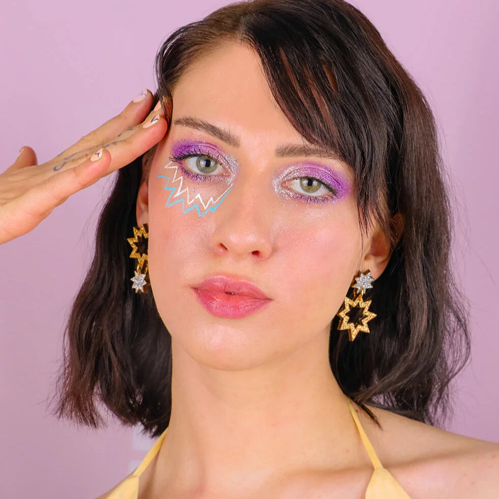 Person with colorful makeup and star-shaped earrings against a pink background