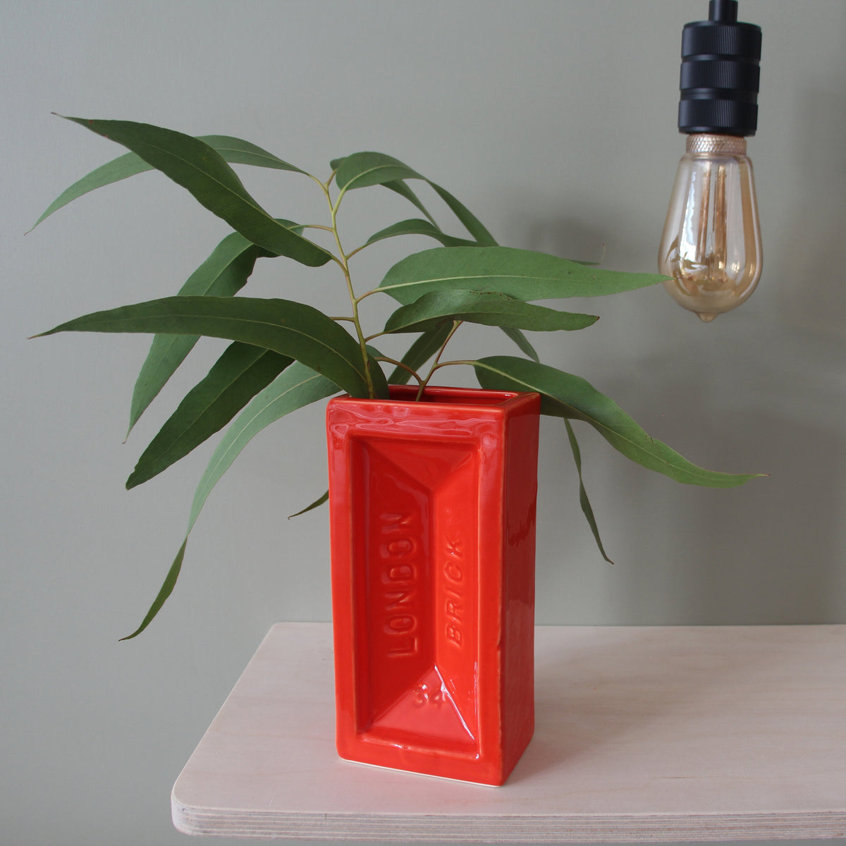 A red ceramic brick-shaped vase on a table with a plant in it.