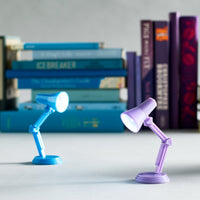 Two mini blue and purple desk lamp shaped booklights on a table in front of stacks of books. 