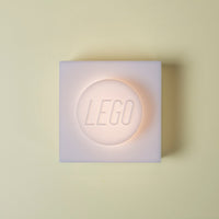 A square LEGO wall light in frosted white, glowing with a warm peach-pink light from within, centered on a pale cream background.