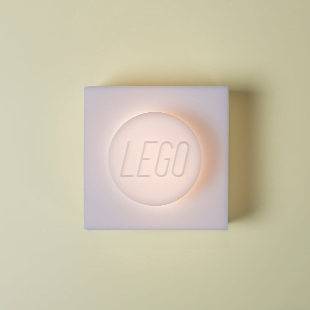A square LEGO wall light in frosted white, glowing with a warm peach-pink light from within, centered on a pale cream background.