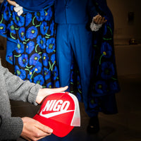 Person holding a red cap with 'NIGO' logo in front of a blue floral dress and suit.
