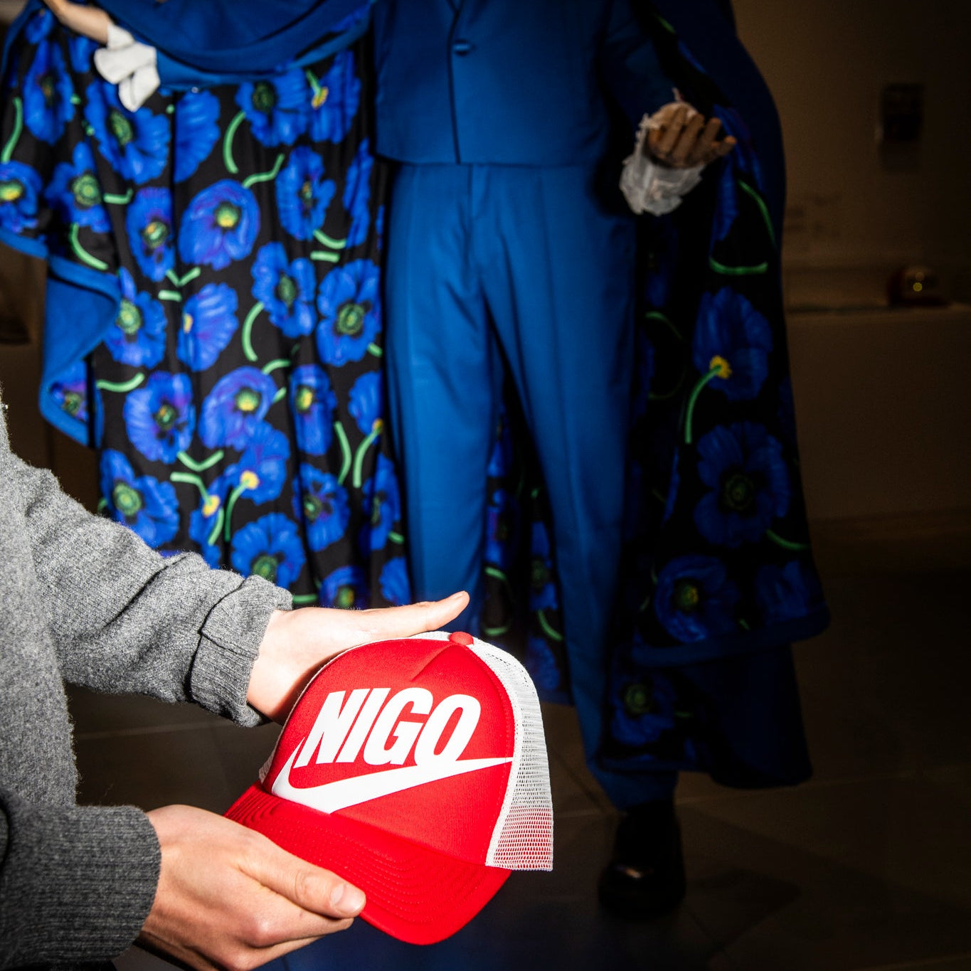 Person holding a red cap with 'NIGO' logo in front of a blue floral dress and suit.