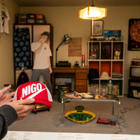 Person holding a red cap with 'NIGO' branding in a room with shelves and decor.
