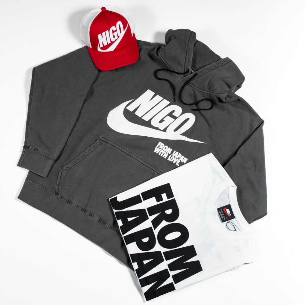 A styled flat-lay of a dark gray Nike hoodie with a large white 'NICO' and swoosh graphic, paired with a red and white Nike cap and a white graphic T-shirt with bold black text reading 'FROM JAPAN'.