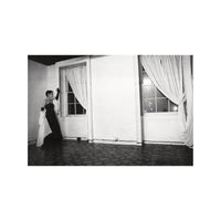 Black and white photo of a person in formal attire standing in a room with large windows and curtains.