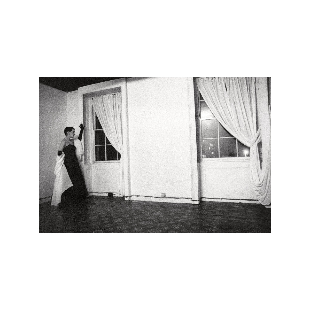 Black and white photo of a person in formal attire standing in a room with large windows and curtains.