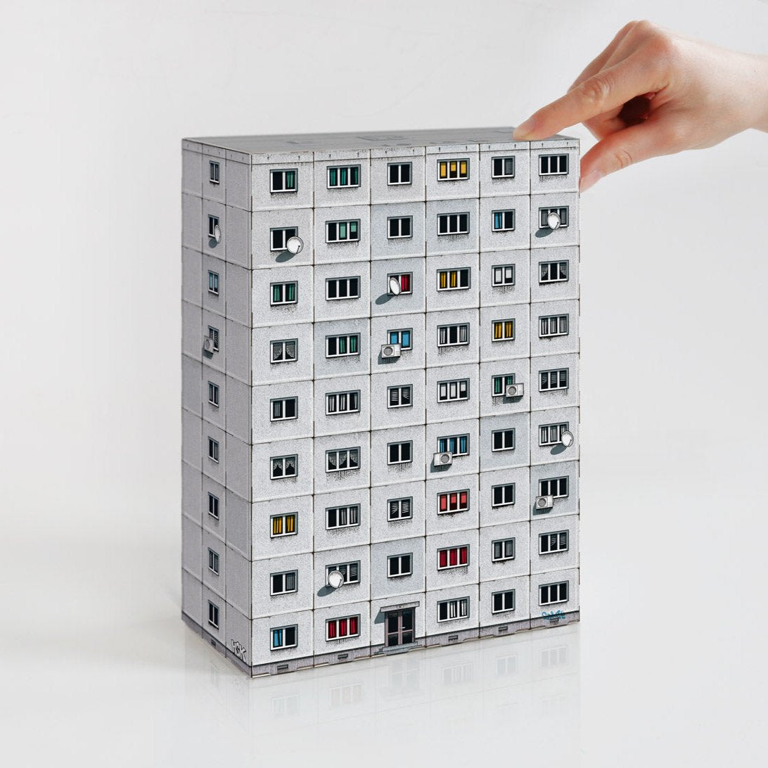 A person's hand putting together a grey paper model of a Brutalist apartment building. 