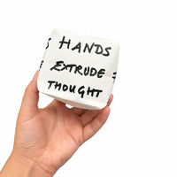 A hand holding a small white ceramic cup with black handwritten text that reads 'HANDS EXTRUDE THOUGHT' in an indoor office or studio setting.