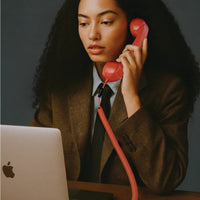 Person in a suit holding a red phone receiver with an Apple laptop on a desk