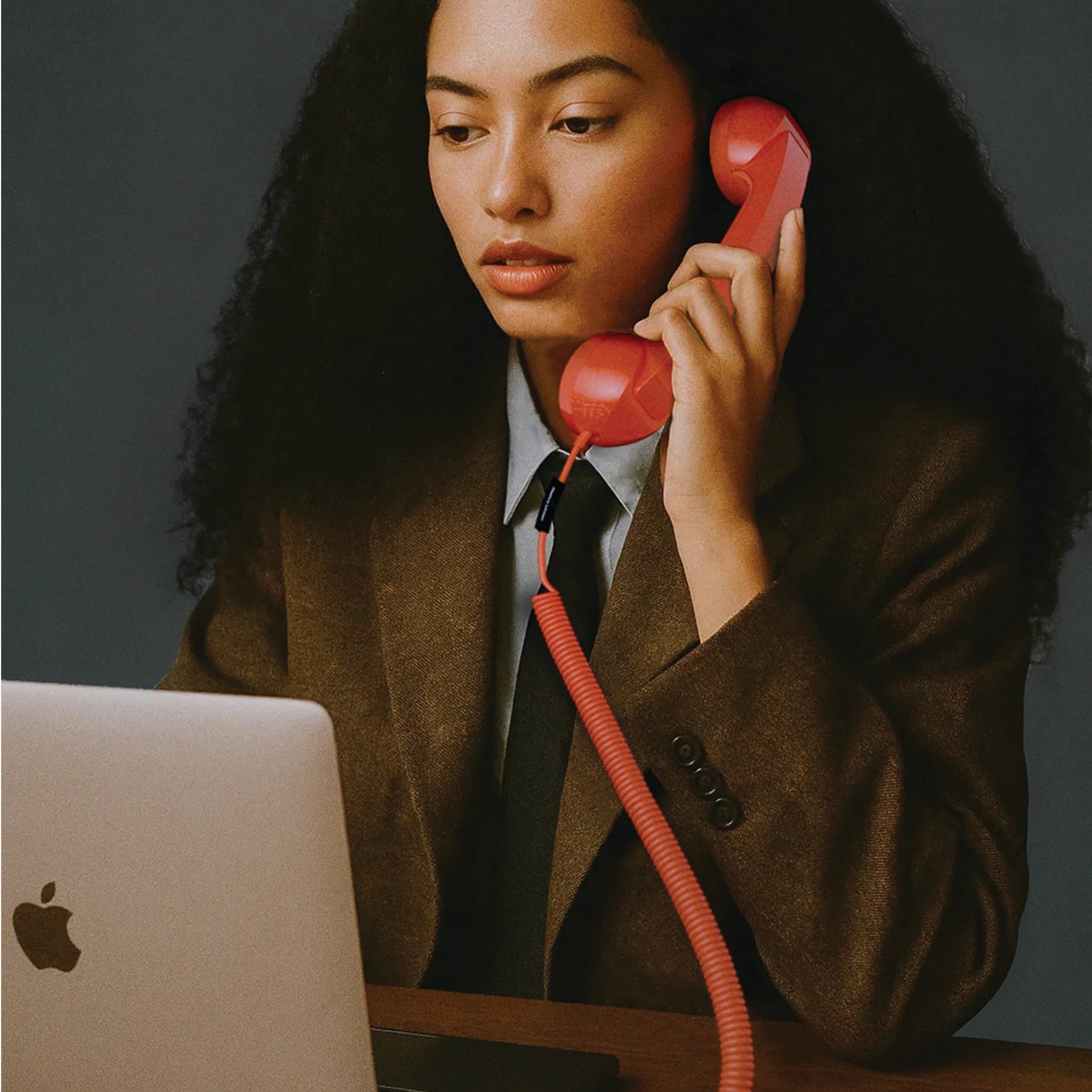 Person in a suit holding a red phone receiver with an Apple laptop on a desk